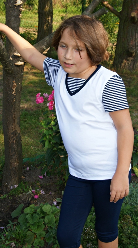 Dziewczynka w koszulce białej z krótkim rękawem, bawełnianej, krótkie rękawy w wąskie paski biało-granatowe, wykończenie pod szyją granatową lamówką i wstawką ozdobną w paski biało-granatowe, fason VKD-51, w stylizacji z granatowymi leginsami.jpg