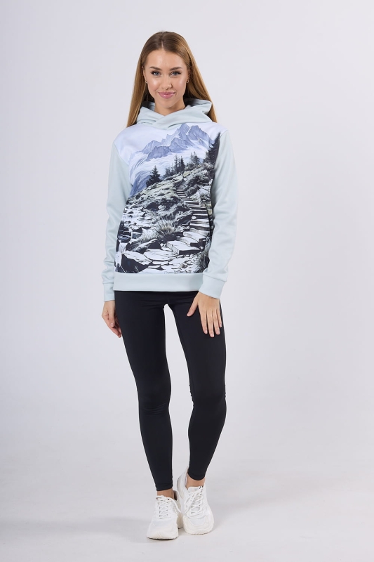 Modelka w bluzie hoodie fason VBL-46 z grafiką Mountains 0055, grafika - szkic przedstawiający góry, las, strumyk, ścieżkę, w odcieniach szarości, bieli, błękitu, bluza z kieszenią typu kangurka, z kapturem, w stylizacji z czarnymi długimi leginsami.jpg