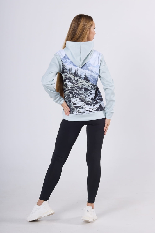 Modelka stojąca tyłem w bluzie hoodie fason VBL-46 z grafiką Mountains 0055, grafika - szkic przedstawiający góry, las, strumyk, ścieżkę, w odcieniach szarości, bieli, błękitu, w stylizacji z czarnymi długimi leginsami.jpg