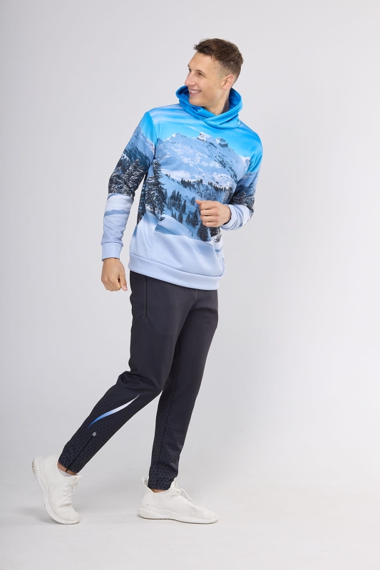Model w bluzie dresowej nierozpinanej z kapturem, kieszeń typu kangurka, wyższa stójka, fason VBL-46 z grafiką Wilk 0111, z przodu ośnieżone góry, z tyłu na tle gór głowa wilka, w stylizacji z czarnymi sportowymi spodniami.jpg