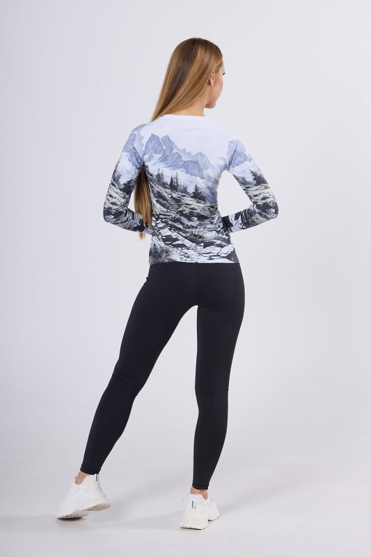 Modelka prezentuje odzież stojąc tyłem longsleeve fason VKDRd-102 grafika Mountains 0055 z czarnymi leginsami, koszulka krój dopasowany, przedstawiający górską scenerię, w kolorystyce dominują odcienie bieli, szarości, czerni i delikatnego błękitu.jpg