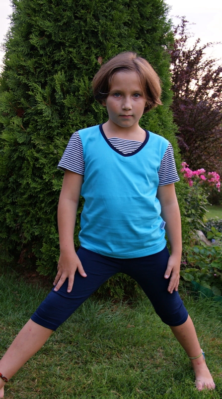 Dziewczynka w koszulce niebieskiej z krótkim rękawem, bawełnianej, krótkie rękawy w wąskie paski biało-granatowe, wykończenie pod szyją granatową lamówką i wstawką ozdobną w paski biało-granatowe, fason VKD-51, w stylizacji z granatowymi leginsami.jpg